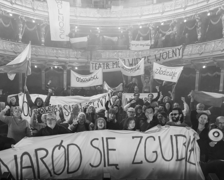 Czarno-białe zdjęcie. Tłum protestujących w przestrzeni widowni dużej sceny teatru. Trzymają transparenty z hasłami: „Naród się zgubił”, „Teatr mój widzę wolny”, „Oszuści”, „Złodzieje”, a także „Wolność”. Na balkonach widoczny jest dodatkowy transparent, „Mam już dość teatru. Część osób unosi pięści, jedna trzyma megafon.